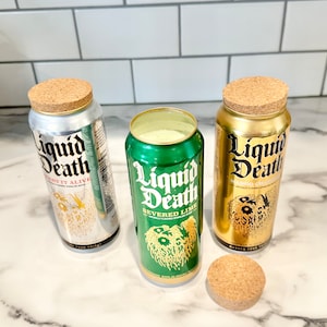 May include: Three aluminum cans with cork lids. The cans are labeled "Liquid Death" with different flavors: "Cherry It Alive", "Severed Lime", and "Mango Chainsaw".