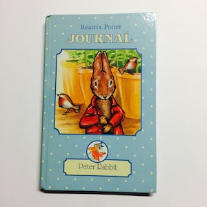 May include: A light blue journal with white polka dots featuring a Peter Rabbit illustration. The cover displays the text "Beatrix Potter JOURNAL" above a picture of Peter Rabbit in a red coat. The journal has a light blue border with the words "Peter Rabbit".