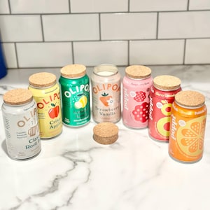 May include: A collection of canned beverages with cork lids. Flavors include Crisp Apple, Lemon Lime, Strawberry Vanilla, Raspberry Rose, Cherry Limeade, and Orange. The cans are arranged on a white surface.