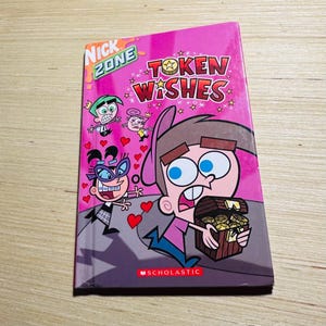 May include: A children's book titled "Token Wishes" with cartoon characters on the cover. The cover is pink with the words "Nick Zone" and "Scholastic" printed on it. The book features a cartoon character holding a treasure chest.