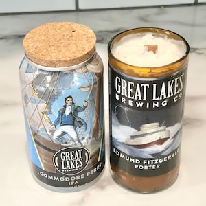 May include: Two repurposed glass containers. One is a jar with a cork lid, featuring a label with a man on a ship and the text "Commodore Perry IPA". The other is a candle with the text "Great Lakes Brewing Co" and "Edmund Fitzgerald Porter".