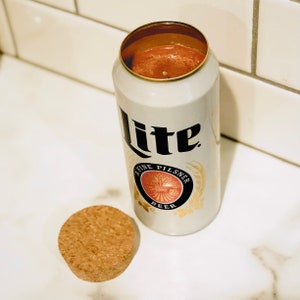 May include: A white aluminum can with a red wax candle inside. The can has the text "Lite" and "A Fine Pilsner Beer" printed on it.
