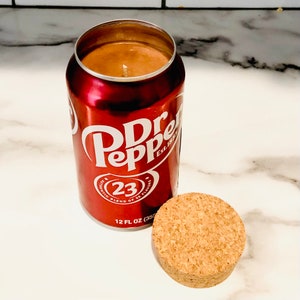 May include: A red and white Dr Pepper can candle with a brown wax top and a cork lid. The can has the text "Dr Pepper" and "Est. 1885" printed on it. The can also has the text "23" and "23 Flavors" printed on it. The can is 12 fl oz (355 ml).