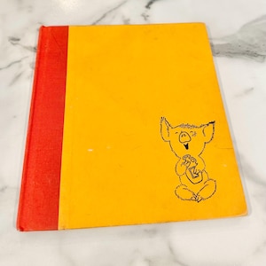 May include: A hardcover book with a bright yellow cover and a red spine. The front cover features a black line drawing of a small, seated koala-like creature holding a bottle. The design is simple, with the illustration positioned on the right side.