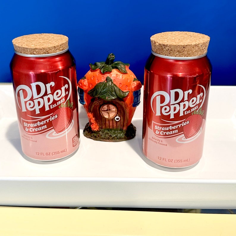 May include: Two 12-ounce cans of Dr Pepper Strawberries & Cream soda. The cans are red with a white label and a pink strawberry graphic. The text "Dr Pepper Est. 1885" is printed on the label.