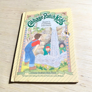 May include: A Cabbage Patch Kids storybook titled Xavier's Fantastic Discovery, featuring a yellow cover with a diagonal stripe pattern. The illustration shows a waterfall, a child in blue overalls, a small figure, and a bee-striped rabbit.