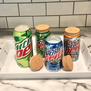 May include: Four Mountain Dew soda can candles with cork lids on a white tray. The cans feature the brand's logo and various color schemes, including green, blue, and red. Two cork lids are placed separately.