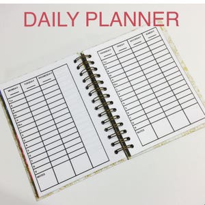 May include: A daily planner with a gold and white floral design. The planner has a spiral binding and is open to show the weekly schedule. The days of the week are listed across the top of the page, and the hours of the day are listed down the side. There is a section for notes at the bottom of each page.