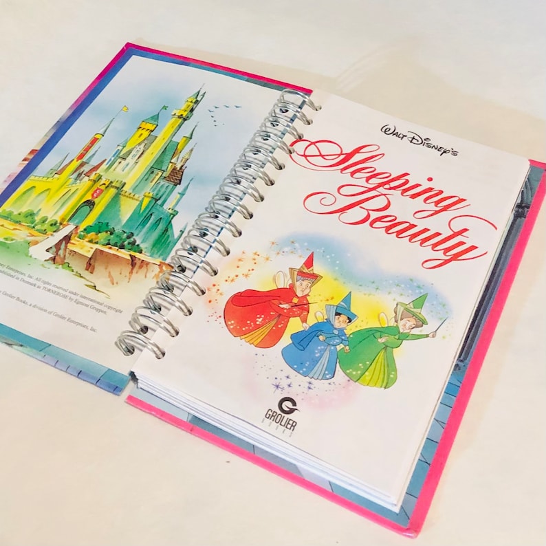 May include: An open book with a spiral binding. The cover features a colourful illustration of Sleeping Beauty's fairies and the text "Walt Disney's Sleeping Beauty". The inside page shows a colourful illustration of Cinderella's castle.