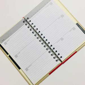 May include: A white daily planner with a black and red striped cover. The planner has a spiral binding and is open to a weekly view. The days of the week are labelled Monday through Sunday. The planner has a section for addresses and notes.
