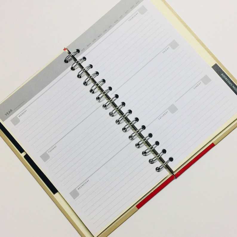 May include: A white daily planner with a black and red striped cover. The planner has a spiral binding and is open to a weekly view. The days of the week are labelled Monday through Sunday. The planner has a section for addresses and notes.