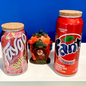May include: Two red and white soda cans, one labeled "Faygo Red Pop" and the other "Fanta Strawberry Flavored Soda". Both cans are on a white surface with a small red and green ceramic strawberry house in the center.