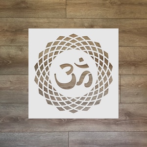 May include: A white stencil featuring the Om symbol within a geometric mandala design. The stencil is square and cut from a solid white material. The background is a wooden surface with brown planks.