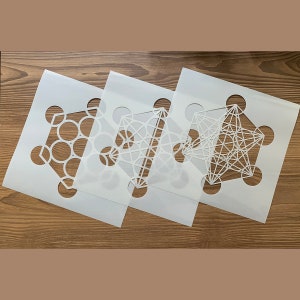May include: Three white stencils with geometric designs. The stencils are layered on top of each other. The designs include a circle pattern, a star pattern, and a flower pattern.
