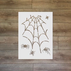 Könnte beinhalten: Eine weiße Schablone mit einem Spinnennetz-Design und vier Spinnen. Die Schablone ist perfekt, um eine gruselige Halloween-Atmosphäre zu schaffen.