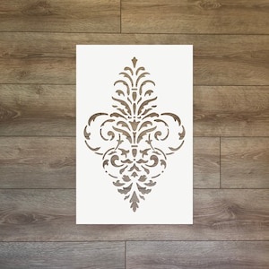 Puede incluir: Un esténcil blanco con un diseño simétrico y ornamentado de elementos florales y de volutas. El esténcil rectangular se coloca sobre un fondo de madera, mostrando el intrincado patrón recortado.