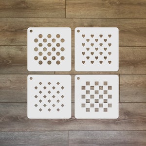 May include: Four white square stencils with different patterns: polka dots, hearts, diamonds, and a checkerboard design. Each stencil has a small hole at the top for hanging. The stencils are displayed on a wooden surface.