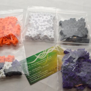 May include: Four bags of colorful plastic building blocks. The bags are clear and show the blocks inside. The blocks are orange, white, black, and purple.