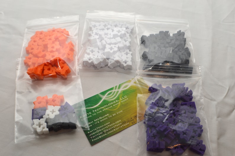 May include: Four bags of colorful plastic building blocks. The bags are clear and show the blocks inside. The blocks are orange, white, black, and purple.