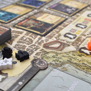 May include: A close-up of a game board with various game pieces, including black and white figures, and orange cubes. The board features a map with a brown and white background. The text "The Tower of Luck" is visible on a card in the lower left corner.