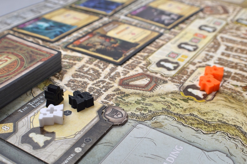 May include: A close-up of a game board with various game pieces, including black and white figures, and orange cubes. The board features a map with a brown and white background. The text "The Tower of Luck" is visible on a card in the lower left corner.
