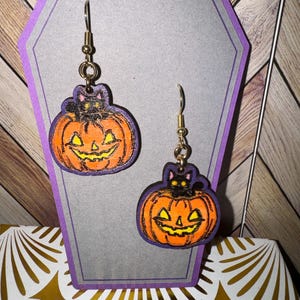 May include: A pair of Halloween-themed earrings featuring a black cat sitting on top of a carved pumpkin. The pumpkins are orange with a black outline and a carved jack-o'-lantern face. The earrings are made of wood and have gold-colored hooks.