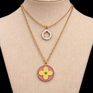 May include: A two-strand necklace set with gold-colored chains. The top necklace has a small white pendant with a clover-shaped design. The bottom necklace has a larger pendant with a pink background, a yellow clover, and a small gem.