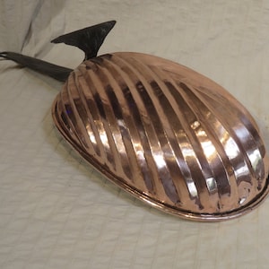 May include: A copper-colored, oval-shaped mold with a ridged design. It has a black handle and a decorative element. The mold is likely used for baking or cooking. The overall design is vintage.