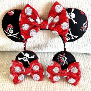 May include: Red and white polka dot bow with black fabric ears featuring a skull and crossbones design. The ears are attached to a black headband.