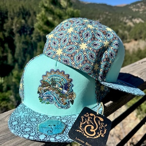 May include: A snapback hat with a turquoise peak and a patterned crown featuring yellow and brown geometric shapes. The hat has a detailed graphic on the front and a tag that reads "Limited Edition".