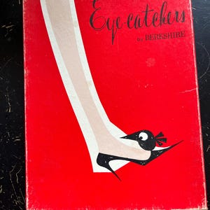 Könnte beinhalten: Eine rote Vintage-Box mit dem Text "Eye catchers by BERKSHIRE" in schwarzer Schrift. Die Box zeigt eine stilisierte Illustration eines Beins und Fußes, der einen schwarzen Schuh mit Vogelmotiv trägt.