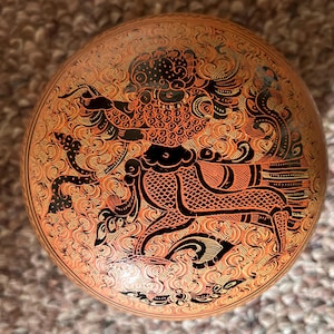 以下が含まれることがあります： 黒とオレンジの複雑なイラストが施された、丸いオレンジ色の装飾ボックス。 デザインは、詳細なパターンと様式化された動物を持つ中心的な人物を特徴とし、文化的な芸術性と職人技を表現しています。