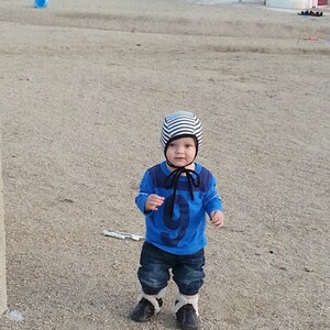 May include: A child in a blue top with the number 9, dark denim jeans, and black boots with white trim. The child is wearing a striped hat with a black tie. The child is standing on a gravel surface.