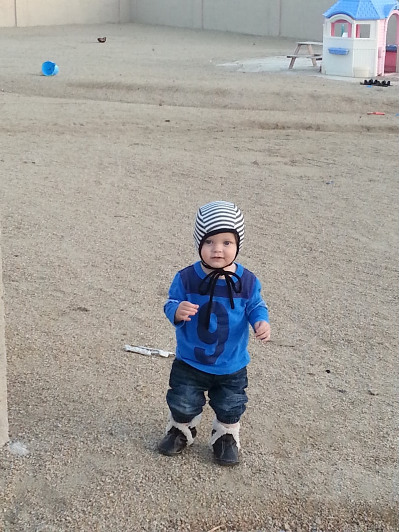 May include: A child in a blue top with the number 9, dark denim jeans, and black boots with white trim. The child is wearing a striped hat with a black tie. The child is standing on a gravel surface.