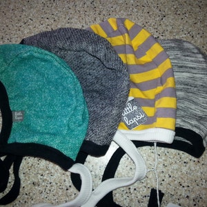 May include: Four baby bonnets in teal, grey, yellow striped, and heather grey. Each bonnet has a black brim and ties. One bonnet has a tag that reads "little lapsi".