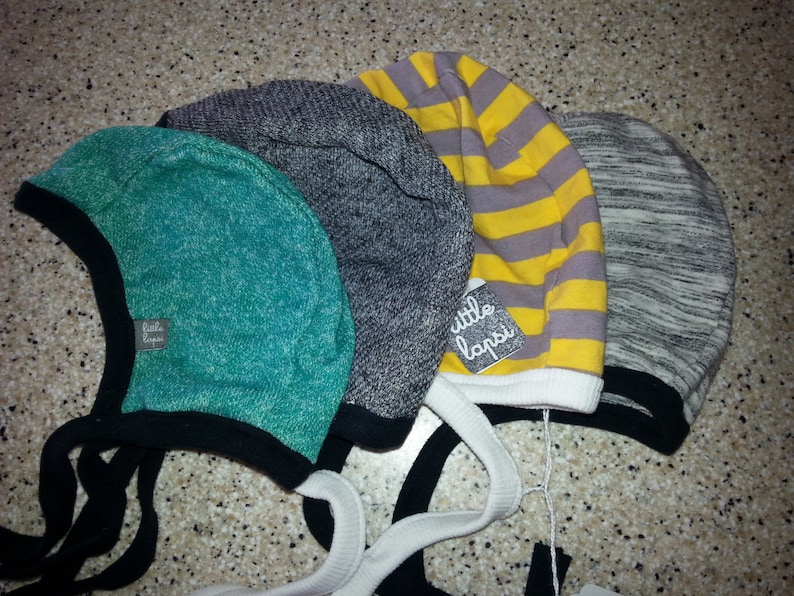 May include: Four baby bonnets in teal, grey, yellow striped, and heather grey. Each bonnet has a black brim and ties. One bonnet has a tag that reads "little lapsi".