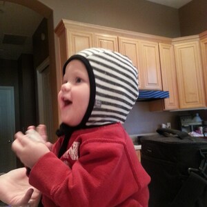 May include: A child in a red top and a black and white striped hat with a black trim. The child is smiling. The background shows a kitchen with wooden cabinets. The hat is a baby accessory.