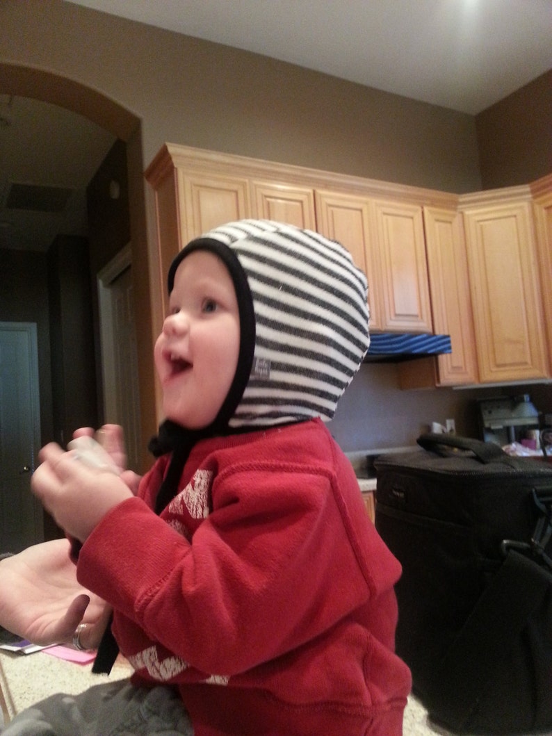 May include: A child in a red top and a black and white striped hat with a black trim. The child is smiling. The background shows a kitchen with wooden cabinets. The hat is a baby accessory.