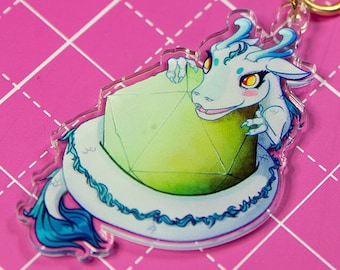 May include: A white dragon with blue accents is curled around a green gemstone. The dragon has pink eyes and a cute expression. The charm is attached to a gold star-shaped keychain.
