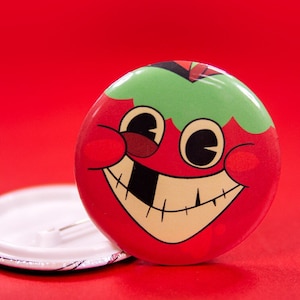 May include: A circular badge featuring a cartoon tomato character. The tomato is red with a green top and a smiling face with a missing tooth. Another badge back is visible.
