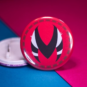 Puede incluir: Un botón rojo, blanco y negro con un diseño estilizado en forma de 'M'. El botón es circular y tiene un acabado brillante.