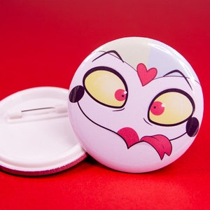 Puede incluir: Un botón blanco con una cara de dibujos animados de una criatura rosa con ojos rojos y amarillos y un corazón rojo en la frente. El botón tiene un alfiler en la parte posterior.
