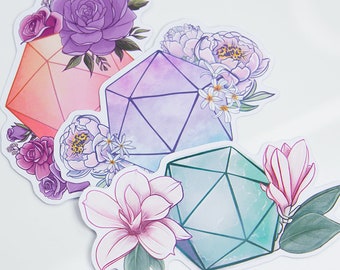 May include: Three geometric shapes, resembling dice, are surrounded by floral designs. The shapes are a pink, a purple, and a blue-green. The flowers are in shades of pink, purple, and white.
