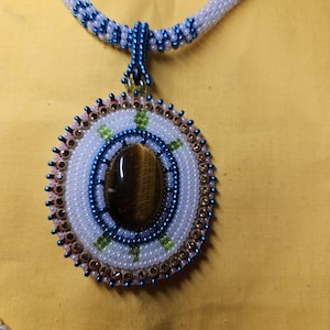 Può includere: Collana di perline fatta a mano con un ciondolo rotondo. Il ciondolo presenta una pietra ovale centrale, circondata da perline bianche, blu, verdi e dorate. Il cordino della collana è bianco con sezioni di perline blu e verdi.