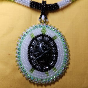 Può includere: Collana di perline fatta a mano con un ciondolo circolare. Il ciondolo ha un centro in pietra nera, circondato da perline bianche, verdi e nere. Il cordino della collana è bianco, nero e verde. Il gioiello è su sfondo giallo.