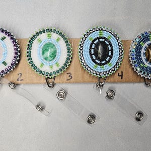 May include: A collection of six beaded badge reels with various gemstone centers and intricate beadwork in shades of green, blue, purple, and white. Each reel has a clear plastic strap and metal clip for attaching to clothing.
