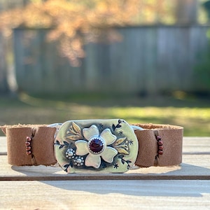 以下が含まれることがあります： 銀色の花の留め金が付いた茶色の革製カフブレスレット。留め金には、赤い宝石の中心と金のアクセントが付いた花があります。革バンドは、小さな濃い赤色のビーズで飾られています。