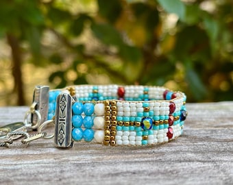 Op de afbeelding: Een kralenarmband met turquoise, witte en gouden kralen. De armband heeft een zilveren sluiting en een geometrisch patroon. Een handgemaakt accessoire.