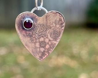 Op de afbeelding: Een hartvormige hanger van koper met een donker, geëtst bloemenpatroon op het oppervlak. Een enkele, ronde, dieprode edelsteen is gezet in een zilverkleurige zetting nabij de linkerbovenhoek. De hanger hangt aan een zilverkleurige ring.