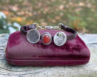 Op de afbeelding: Een donkerbruine leren armband met drie zilverkleurige bedels: een ronde bedel met een gegraveerde eekhoorn, een centrale ovale bedel met een gladde oranje steen en een ronde bedel met een gegraveerde uil en een kleine groene steen.